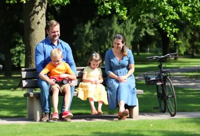 Familie im Stadtpark auf Picknickbank: Vater mit Sohn, Mutter mit Tochter, umgeben von Eichenbäumen und Fahrrad